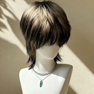 Op de afbeelding: Een mannequinhoofd met een gelaagde, tweekleurige pruik in zwart, bruin en blond. De mannequin draagt een zilveren ketting met een teal-kleurige veervormige hanger. De achtergrond is een neutraal beige met schaduwen.