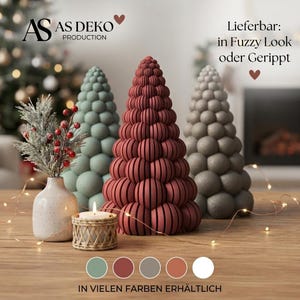 Op de afbeelding: Drie kegelvormige decoratieve bomen met gestructureerde oppervlakken in saliegroen, donkerrood en grijs. Een kleine keramische vaas met kunstgroen en rode bessen staat naast een kaars in een gevlochten houder. Tekst: AS DEKO PRODUCTION.