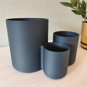 Pu&ograve; includere: Un organizer da scrivania grigio scuro con tre scomparti cilindrici di diverse altezze e larghezze, collegati tra loro. Il contenitore con finitura opaca presenta un design esterno liscio e arrotondato con aperture sulla parte superiore.