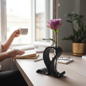 May include: A single lavender rose in a clear glass test tube held by a matte black, curved, sculptural stand. The stand features a fluid, ribbon-like design that supports the upright glass vessel, creating a contrast with the soft floral bloom.