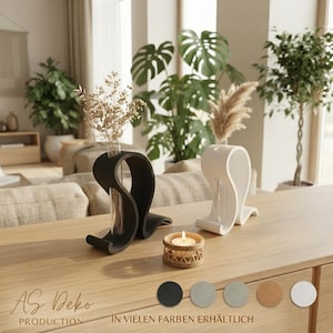 May include: Two curved, wave-shaped vases, one black and one white, each holding a glass tube with dried botanical stems. A small, round, woven candle holder with a lit candle sits between them. Text reads AS Deko Production.