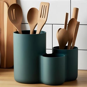Pu&ograve; includere: Un portautensili da cucina color verde ottanio scuro con tre scomparti cilindrici di altezze diverse. Il supporto contiene diversi utensili da cucina in legno chiaro, tra cui cucchiai e una spatola forata.
