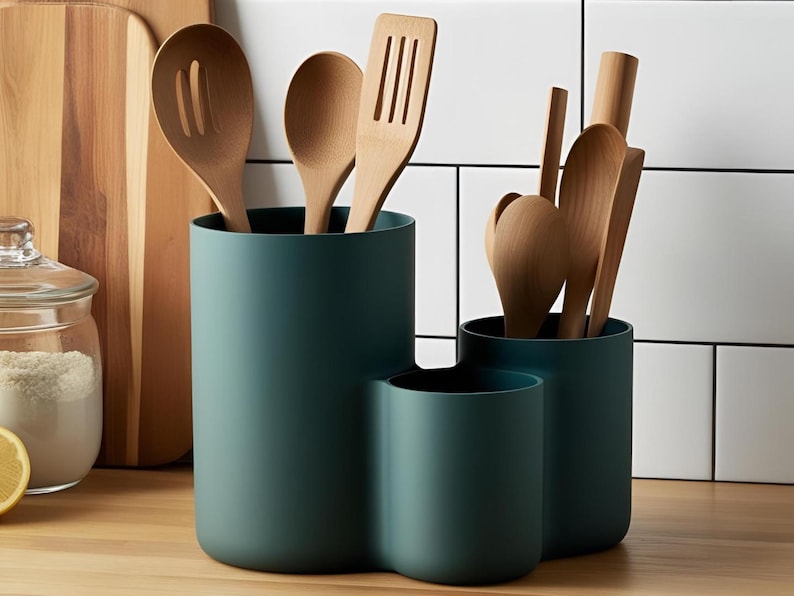 Pu&ograve; includere: Un portautensili da cucina color verde ottanio scuro con tre scomparti cilindrici di altezze diverse. Il supporto contiene diversi utensili da cucina in legno chiaro, tra cui cucchiai e una spatola forata.