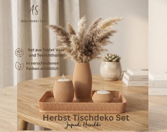 May include: A tan decorative set featuring a rectangular tray, a tall vase, and two small candle holders, all with a consistent wavy, ribbed texture. The vase contains dried pampas grass. Text includes AS DEKO PRODUCTION and Herbst Tischdeko Set.