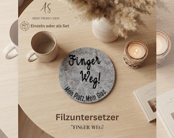 May include: A circular grey felt coaster featuring the black printed text 'Finger Weg! Mein Platz, Mein Glas'. The coaster is positioned on a light-colored wooden surface alongside a ceramic mug, a lit candle in a woven holder, and a book.