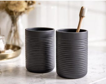 May include: Two cylindrical dark gray bathroom tumblers featuring a horizontal ribbed texture on the exterior. One tumbler contains a single bamboo toothbrush with white bristles, while the other tumbler remains empty.