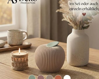May include: A ribbed, apple-shaped decorative object in a light beige tone with a sage green leaf accent. Beside it sits a candle in a jute-wrapped glass holder, a speckled beige vase with dried grasses, and a mug on a wooden tray.