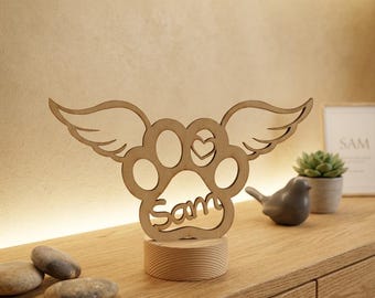 May include: A light-colored wooden cutout featuring a paw print design with angel wings extending from the sides. The center of the paw print includes a small heart cutout, and the name Sam is engraved at the bottom. It stands on a round wooden base.