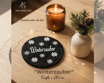 May include: A round black felt coaster featuring white snowflake patterns and the German text Winterzauber in a script font. The coaster is 10 cm in diameter and is displayed alongside a lit candle in a glass jar and a small white vase.