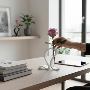 May include: A white, curved, abstract-shaped vase holding a single lavender rose in a slender glass tube. The vase features a smooth, continuous loop design that supports the central stem, creating a clean and modern display piece.