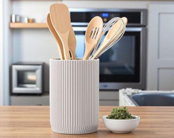 Può includere: Un portautensili cilindrico grigio chiaro con una texture a coste verticali, contenente diversi utensili da cucina in legno chiaro tra cui spatole, un cucchiaio forato e una frusta in metallo, accanto a una piccola ciotola bianca con erbe verdi.