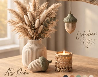 May include: A ribbed beige ceramic vase filled with dried pampas grass and eucalyptus, accompanied by two acorn-shaped ornaments in beige and sage green, and a candle in a woven rattan holder. Text reads: Lieferbar: LIEGEND & HÄNGEND.