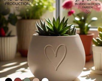 May include: A matte white ceramic planter with a rounded shape and an embossed heart design on the front. The planter contains a green succulent plant. Text on the image reads AS Deko Production, Lieferbar: in verschiedenen Farben, and In verschiedenen Farben erhältlich.