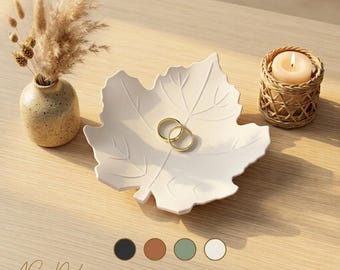 May include: A white leaf-shaped ceramic trinket dish holding two gold rings, accompanied by a small speckled vase with dried grasses and a candle in a woven holder. Text at the bottom reads AS Deko Production and In vielen Farben erhältlich.