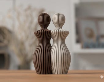 May include: Two abstract human-shaped figurines standing side by side. One figurine is dark brown and the other is off-white. Both feature a vertical ribbed texture with a rounded head, narrow neck, and a curved, hourglass-like body shape.