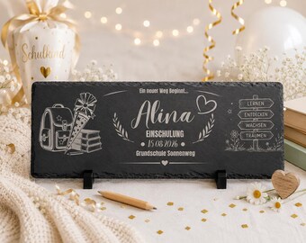 May include: A rectangular black slate plaque featuring white laser-engraved text and graphics. The design includes a school backpack, a cone, books, and a signpost with the words Lernen, Entdecken, Wachsen, Träumen. It displays the name Alina, Einschulung, 15.08.2026, and Grundschule Sonnenweg.
