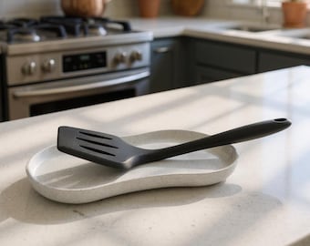 Può includere: Una spatola da cucina nera con fessure appoggiata su un poggia cucchiaio ovale, di colore grigio chiaro e maculato. La spatola presenta un manico lungo con un foro all'estremità e il poggia cucchiaio ha un design leggermente curvo e basso.