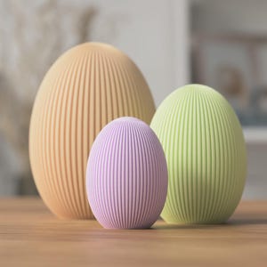 以下が含まれることがあります： 垂直なリブ状のテクスチャを持つ3つの卵型のオブジェクト。一番大きいものはピーチ色、中くらいのものはライトグリーン、一番小さいものはラベンダー色です。各オブジェクトは滑らかで丸みを帯びた上部と平らな底面を持ち、直立しています。