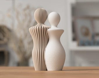 May include: Two abstract sculptures standing side by side. The left sculpture is light beige with a series of vertical, parallel slats. The right sculpture is solid white with a textured, granular surface. Both figures have a rounded head and torso.