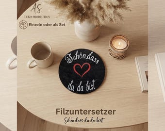 May include: A round black felt coaster featuring a red heart graphic and the white text "Schön, dass du da bist" written in a cursive script. The coaster is positioned on a light wood surface alongside a beige ceramic mug and a candle.