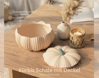 May include: A beige pumpkin-shaped bowl with a matching lid featuring a green stem. The bowl has a ribbed exterior texture. Next to it sits a small white vase with dried pampas grass and a candle in a woven rattan holder on a wooden surface.