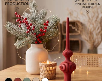May include: A matte red ribbed decorative finial ornament stands next to a white ceramic vase containing frosted pine branches with red berries and a small woven candle holder with a lit candle. Text reads AS Deko PRODUCTION.
