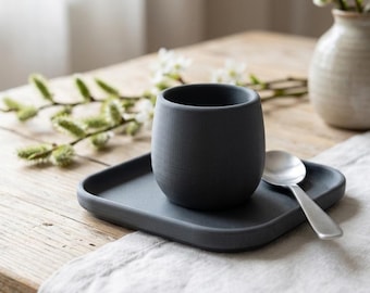 Può includere: Una tazza in ceramica grigio scuro opaco è posizionata al centro di un piattino quadrato abbinato con angoli arrotondati. Un piccolo cucchiaio in metallo color argento poggia sul piattino accanto alla tazza. Il set ha una finitura liscia.