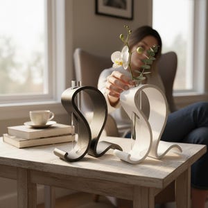 May include: Two curved, S-shaped stands, one matte black and one matte white, each holding a clear glass test tube vase. The white stand contains a single white orchid flower and a green eucalyptus sprig, creating a pair of modern floral displays.