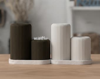 May include: A set of four cylindrical containers with vertical ribbed textures, arranged on two white oval trays. Two containers are dark brown and two are white. One dark brown container holds a cluster of white cotton swabs.