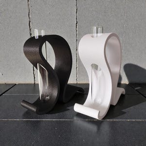 May include: Two curved, S-shaped stands, one matte black and one matte white, each holding a clear glass test tube. The stands have a textured surface and a smooth, continuous loop design that supports the vertical glass tube in the center.