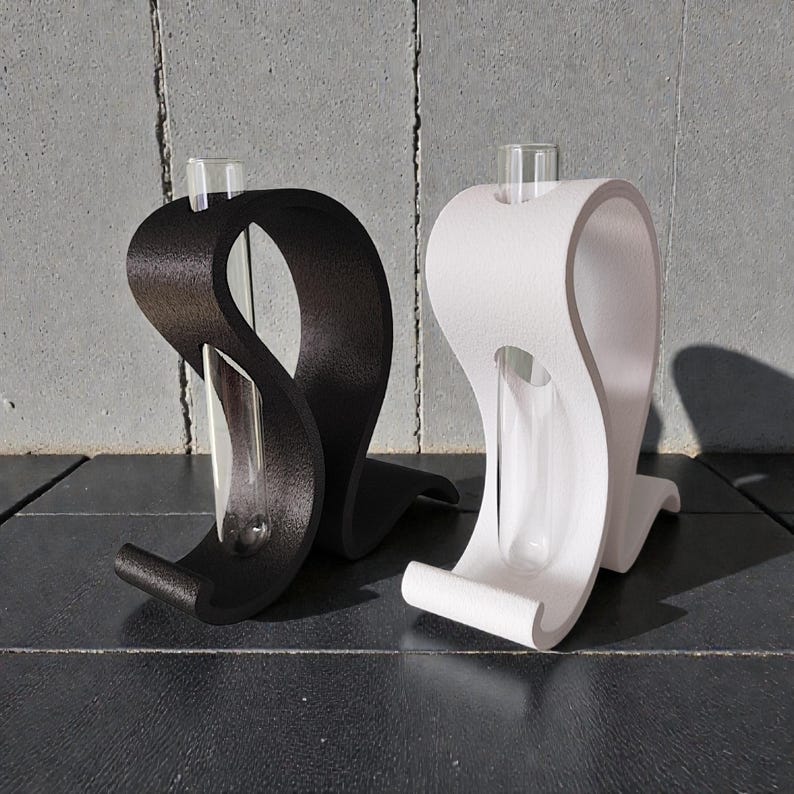 May include: Two curved, S-shaped stands, one matte black and one matte white, each holding a clear glass test tube. The stands have a textured surface and a smooth, continuous loop design that supports the vertical glass tube in the center.