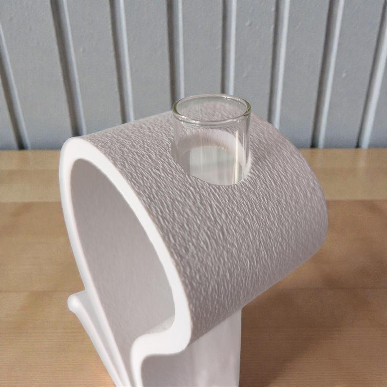 May include: A white, textured, roll-shaped object resembling a toilet paper roll, featuring a smooth, cylindrical glass test tube inserted into the top center. The exterior surface has a matte, granular finish with a curved, open design.