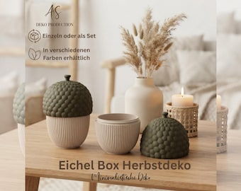 May include: Two acorn-shaped containers with textured olive green lids and ribbed cream bases, one with the lid on and one off. Beside them are a cream vase with dried grasses, a candle in a woven holder, and text reading AS DEKO PRODUCTION.