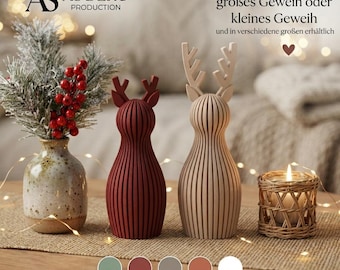 Può includere: Due statuette di renna verticali a coste, una rosso scuro e una beige, accanto a un vaso in ceramica maculato con bacche rosse e un portacandele intrecciato. Testo: AS DEKO PRODUCTION e IN VIELEN FARBEN ERHÄLTLICH.