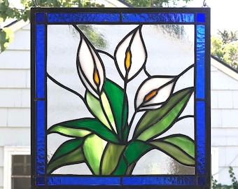 May include: A stained glass window featuring three white calla lilies with green leaves, framed in a dark blue border.