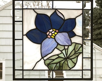 May include: A stained glass window featuring a blue and purple flower with green leaves. The flower is surrounded by a black frame.