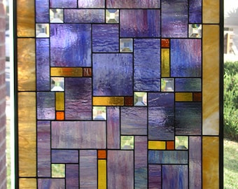 May include: A stained glass window with a geometric pattern of purple, yellow, and orange glass pieces. The window is framed in black and has a gold border.