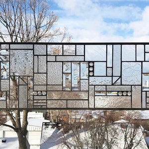 May include: Rectangular stained glass panel with a geometric design. The panel features clear glass pieces in various shapes and textures, framed by black metal. It hangs from a chain against a backdrop of a winter sky and rooftops.