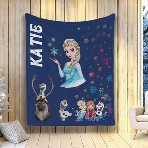 May include: A navy blue blanket with the name "KATIE" in white vertical text. The blanket features illustrations of Elsa, Olaf, and other characters from the film Frozen, along with colourful snowflakes. The blanket is displayed indoors.