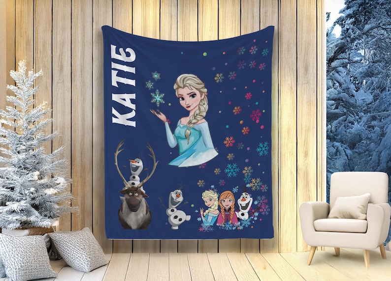 May include: A navy blue blanket with the name "KATIE" in white vertical text. The blanket features illustrations of Elsa, Olaf, and other characters from the film Frozen, along with colourful snowflakes. The blanket is displayed indoors.