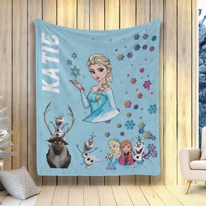 May include: A light blue blanket featuring Elsa from Frozen, with the name "KATIE" in white vertical text. The blanket includes illustrations of Olaf, Sven, and other characters, along with snowflake designs.