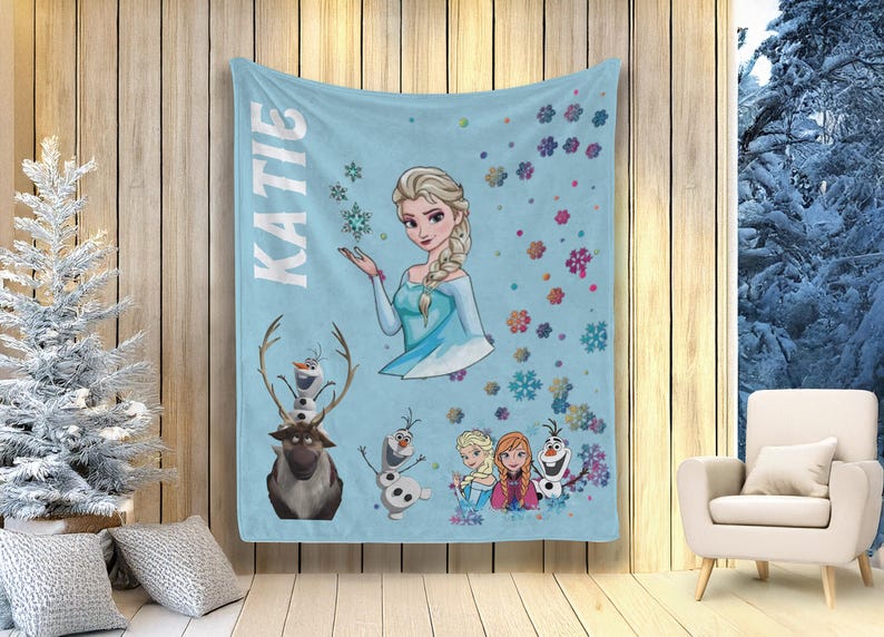 May include: A light blue blanket featuring Elsa from Frozen, with the name "KATIE" in white vertical text. The blanket includes illustrations of Olaf, Sven, and other characters, along with snowflake designs.