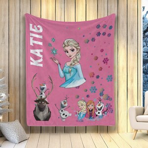 May include: A pink fleece blanket with the name "KATIE" in white, and illustrations of Elsa, Olaf, Anna, and snowflakes. The blanket has a winter theme and is suitable for a child's room.