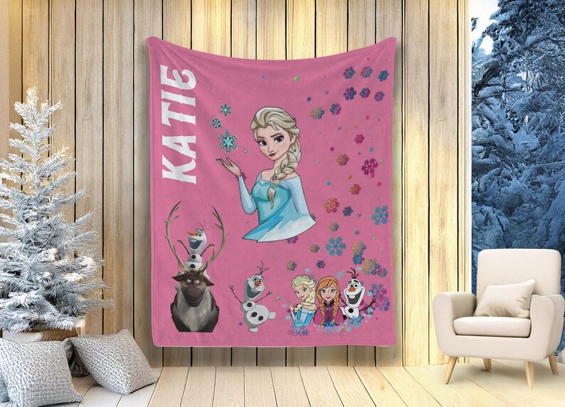 May include: A pink fleece blanket with the name "KATIE" in white, and illustrations of Elsa, Olaf, Anna, and snowflakes. The blanket has a winter theme and is suitable for a child's room.