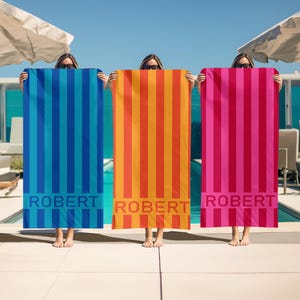May include: Three beach towels with a striped pattern in blue, orange, and pink. Each towel has the name "ROBERT" printed on the bottom.