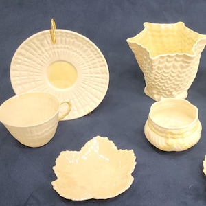 May include: A cream-colored tea set with textured details. The set includes two teacups, two saucers, two leaf-shaped dishes, a small lidded container, and a decorative vase. The pieces are arranged on a dark blue surface.