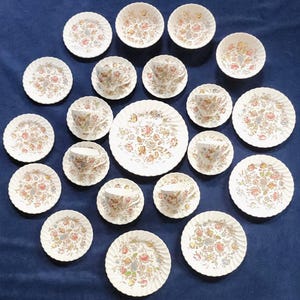 May include: A collection of vintage white porcelain teacups, saucers, bowls, and plates. Each piece features a floral pattern in shades of pink, yellow, and green. The plates have a scalloped edge. The arrangement is on a dark blue surface.