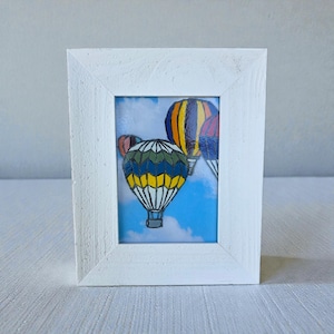 May include: A framed art print featuring three colorful hot air balloons against a blue sky with clouds. The balloons have yellow, blue, green, and red patterns. The white frame has a textured finish.