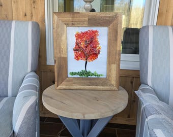 May include: A framed artwork featuring a stylized tree with vibrant red and orange leaves, set against a light background. The tree sits atop a green base, all within a natural wood frame. The artwork is displayed on a round wooden table.