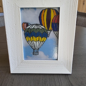 May include: A framed artwork featuring colorful hot air balloons against a blue sky with clouds. The balloons are in shades of yellow, blue, orange, and red. The frame is white with a textured finish.